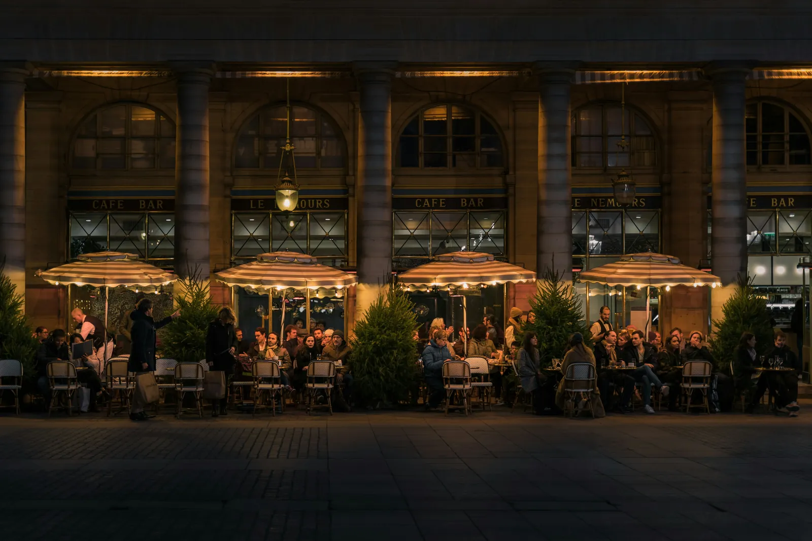 A busy café terrace in the evening, tables full under lit umbrellas